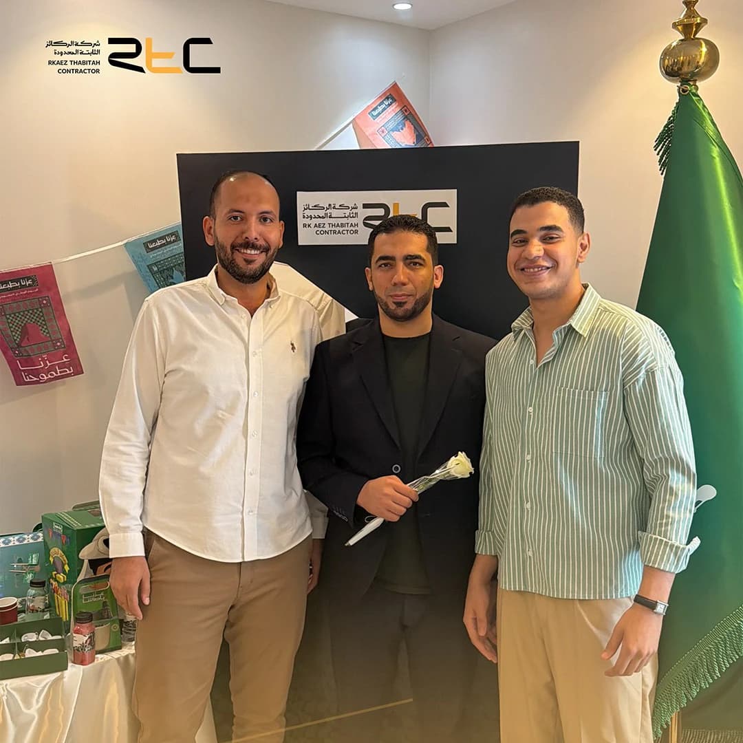 ثلاثة من الحضور أمام خلفية الاحتفال إلى جانب العلم السعودي وطاولة الهدايا، ويحمل أوسطهم وردة بيضاء