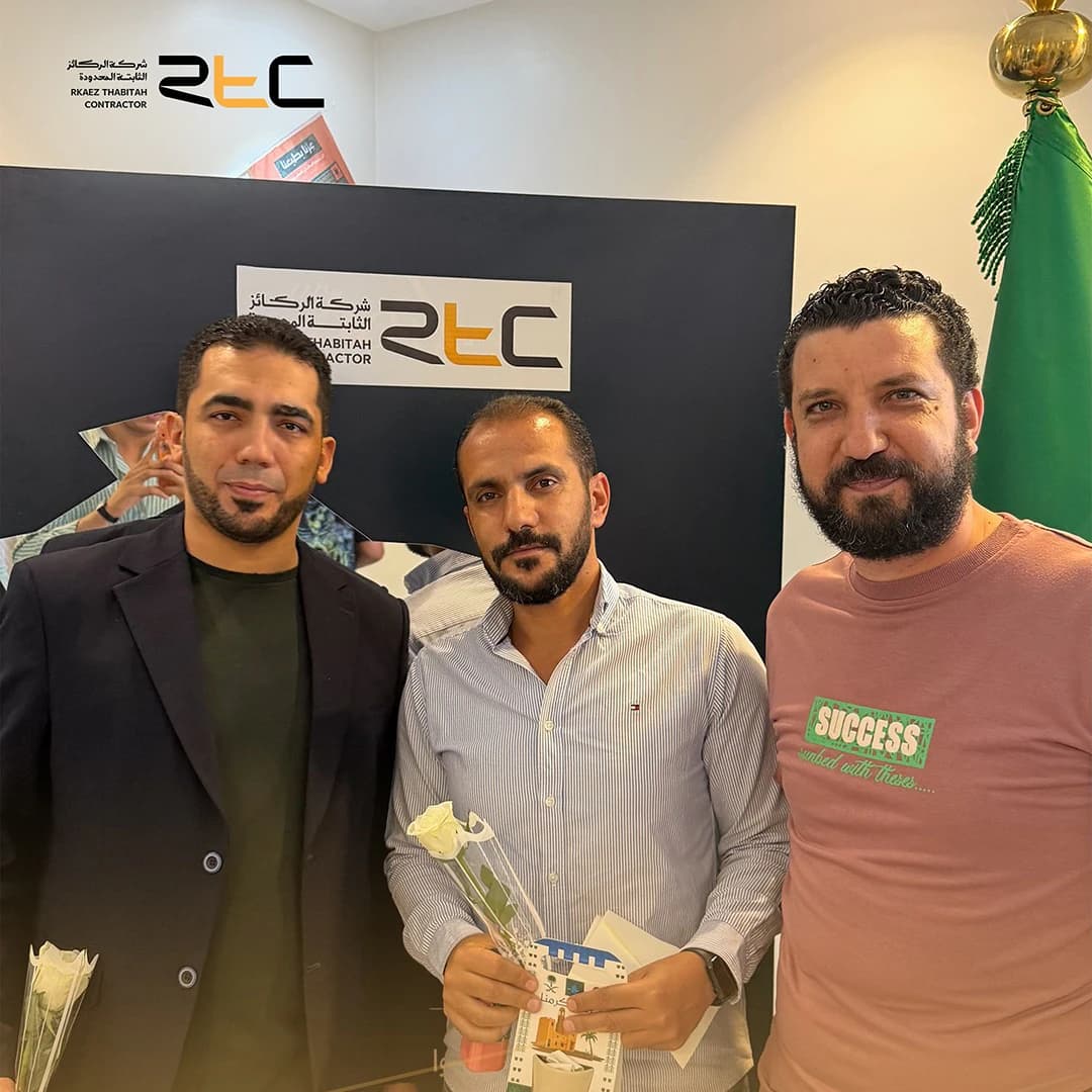 ثلاثة من الحضور أمام خلفية الشركة، ويحمل اثنان منهم ورودًا بيضاء وأحدهما بطاقة هدية