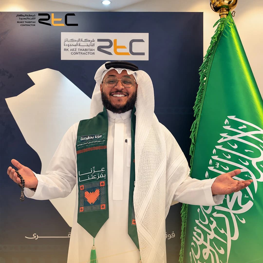 أحد الحضور بالثوب والشماغ الأبيض ووشاح اليوم الوطني، يفتح ذراعيه إلى جانب العلم السعودي