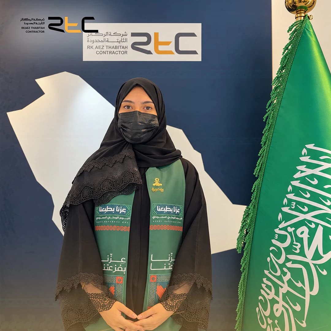 أحد الحضور بعباءة سوداء مزينة بالدانتيل ووشاح اليوم الوطني أمام خلفية الاحتفال