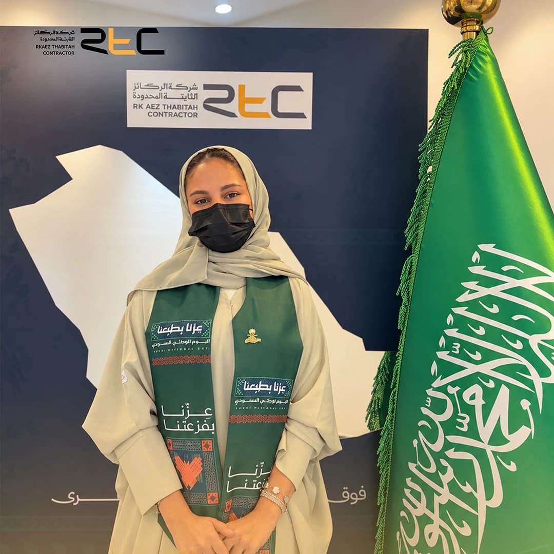 أحد الحضور بعباءة بلون البيج ووشاح اليوم الوطني إلى جانب العلم السعودي