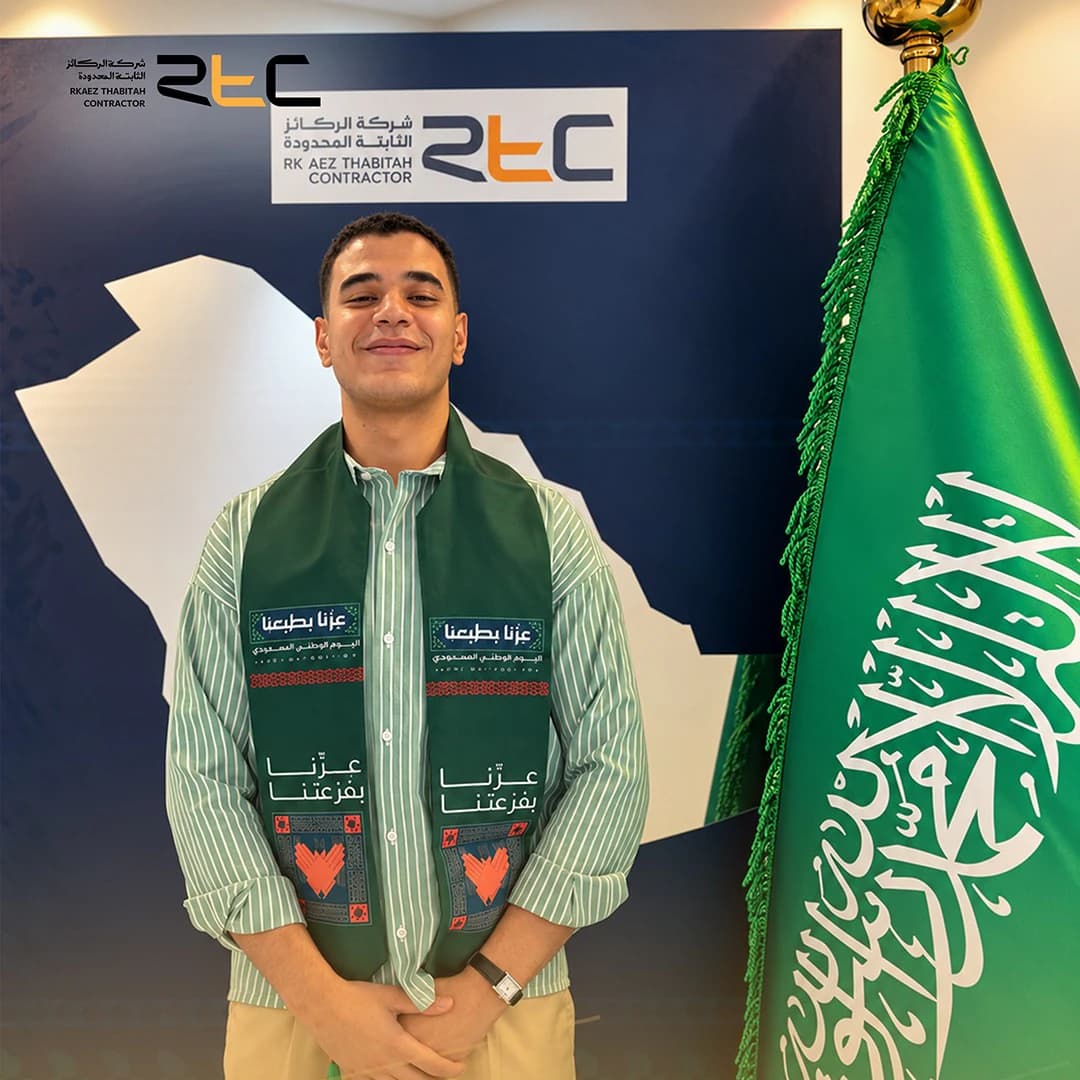 أحد الحضور بقميص مخطط باللون الأخضر ووشاح اليوم الوطني إلى جانب العلم السعودي