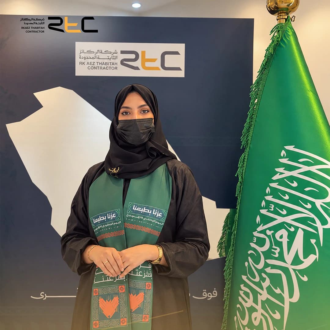 أحد الحضور بعباءة سوداء ووشاح اليوم الوطني، مشبك اليدين إلى جانب العلم السعودي