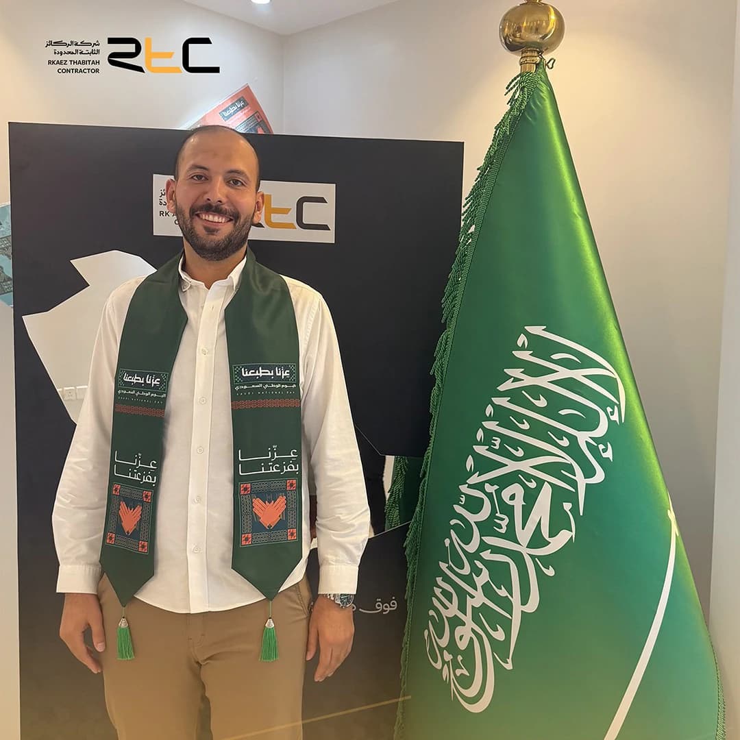 أحد الحضور بقميص أبيض ووشاح اليوم الوطني إلى جانب العلم السعودي