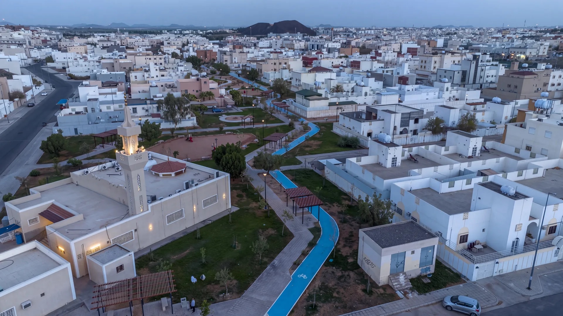 منظر جوي وقت الغروب لممشى الإسكان في حي مهزور ومساره الأزرق الممتد بجوار المسجد والمساحات المزروعة والمظلات